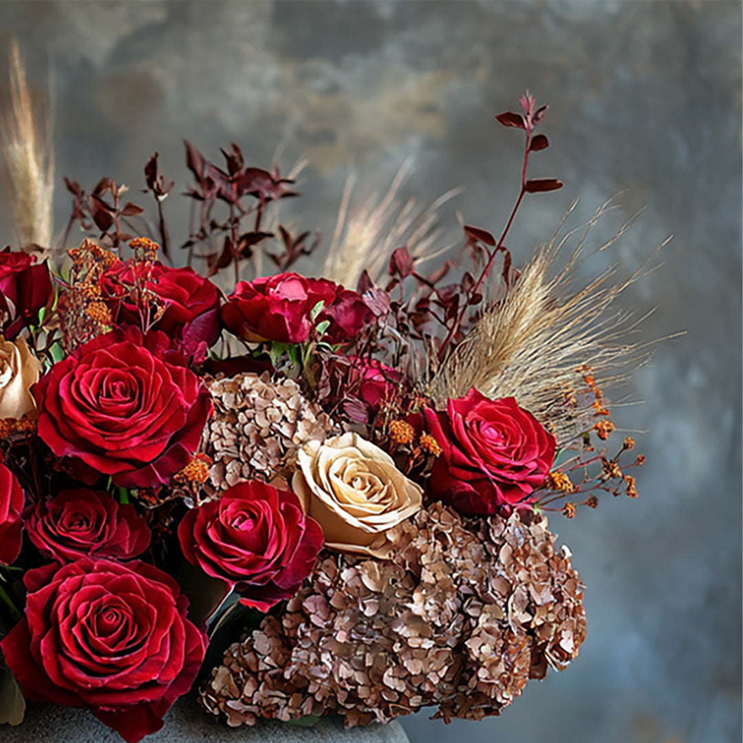 A stunning arrangement of deep red roses, a soft cream rose, and delicate dried florals, arranged artistically with elegant pampas grass and textured elements, creating a rich and captivating composition. This design evokes passion and sophistication, making it perfect for a romantic gesture or a special event.