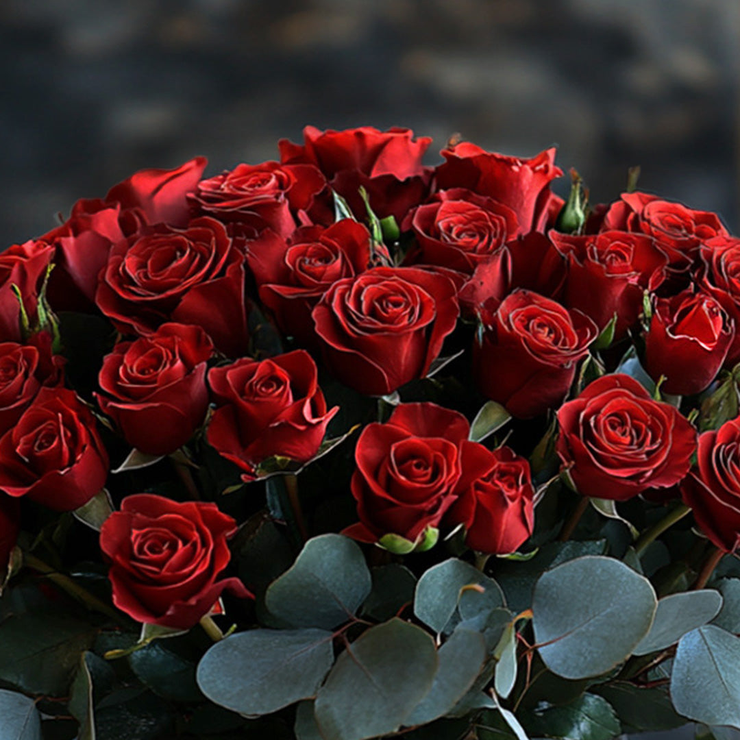 A stunning close-up of a bouquet featuring deep red roses arranged in a lush cluster. The vibrant roses are complemented by soft eucalyptus leaves, creating an elegant, romantic display perfect for special occasions or gifting.