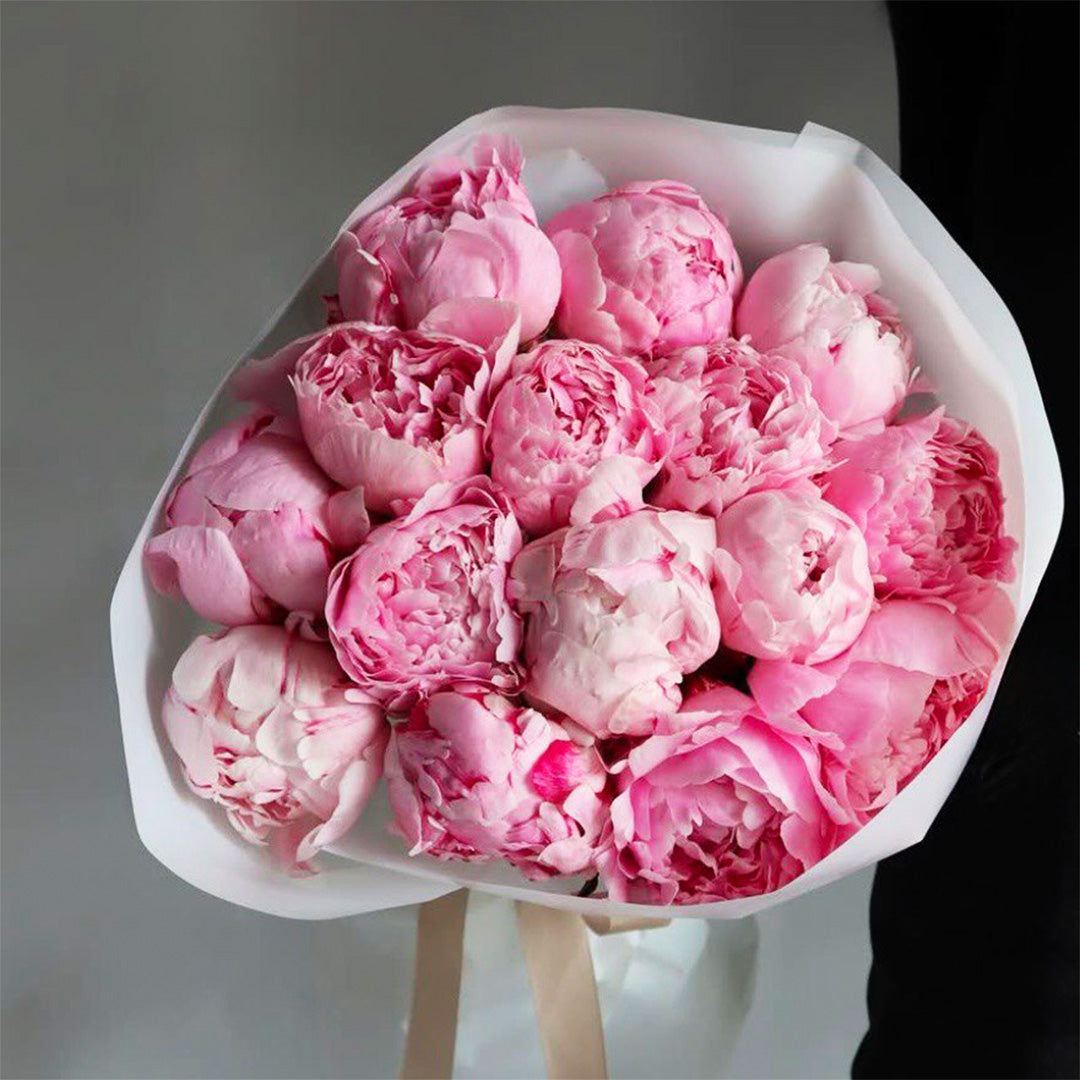 A bouquet of fresh pink peonies, arranged in a soft, rounded shape and wrapped in white paper. The flowers are full-bloom with delicate ruffled petals in various shades of pink, showcasing their natural beauty and elegance.