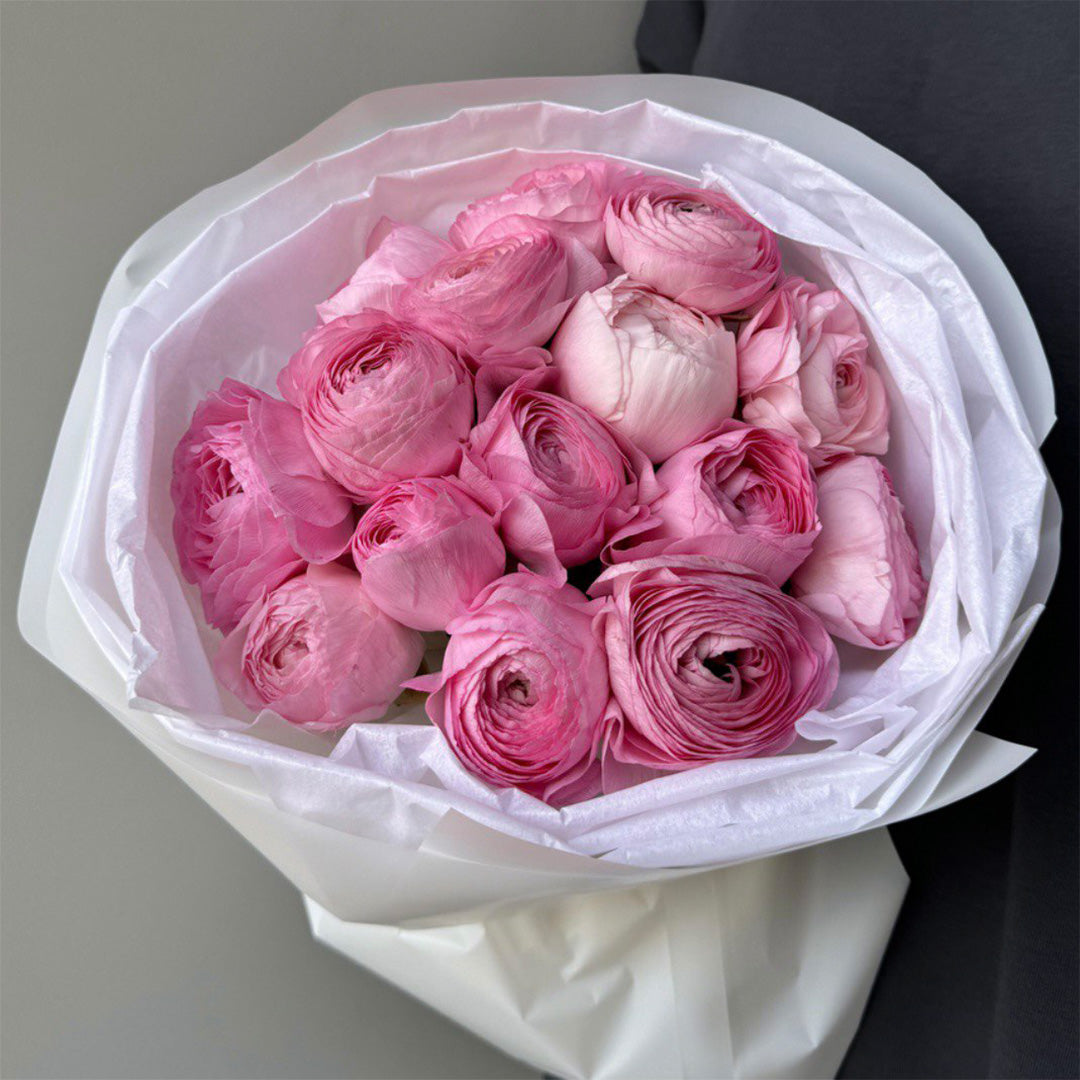 A bouquet of pink ranunculus flowers, their petals delicately layered in soft shades of pink and white. The flowers are tightly packed in the bouquet, wrapped in elegant white paper, creating a lush and full appearance.
