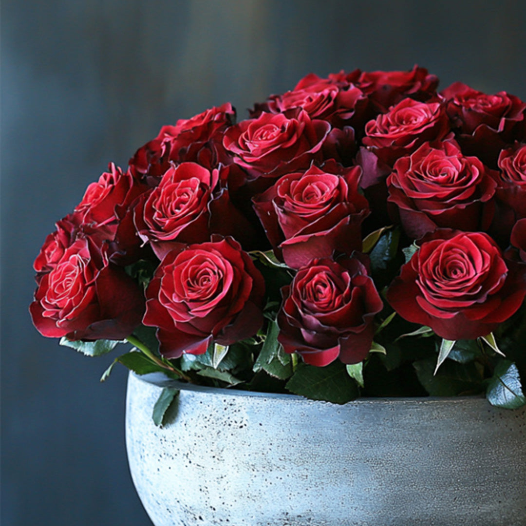 A bouquet of rich, velvety red roses arranged beautifully in a contemporary textured vase, surrounded by lush green leaves, creating an elegant and romantic display.