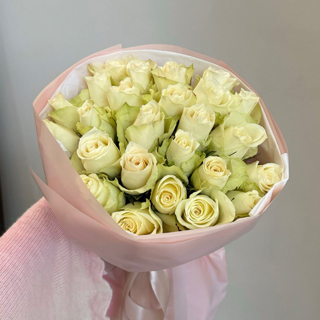 A bouquet of fresh white roses arranged elegantly with soft green foliage. The roses are tightly packed together and wrapped in a soft pink paper with a satin ribbon, creating a graceful and romantic presentation.