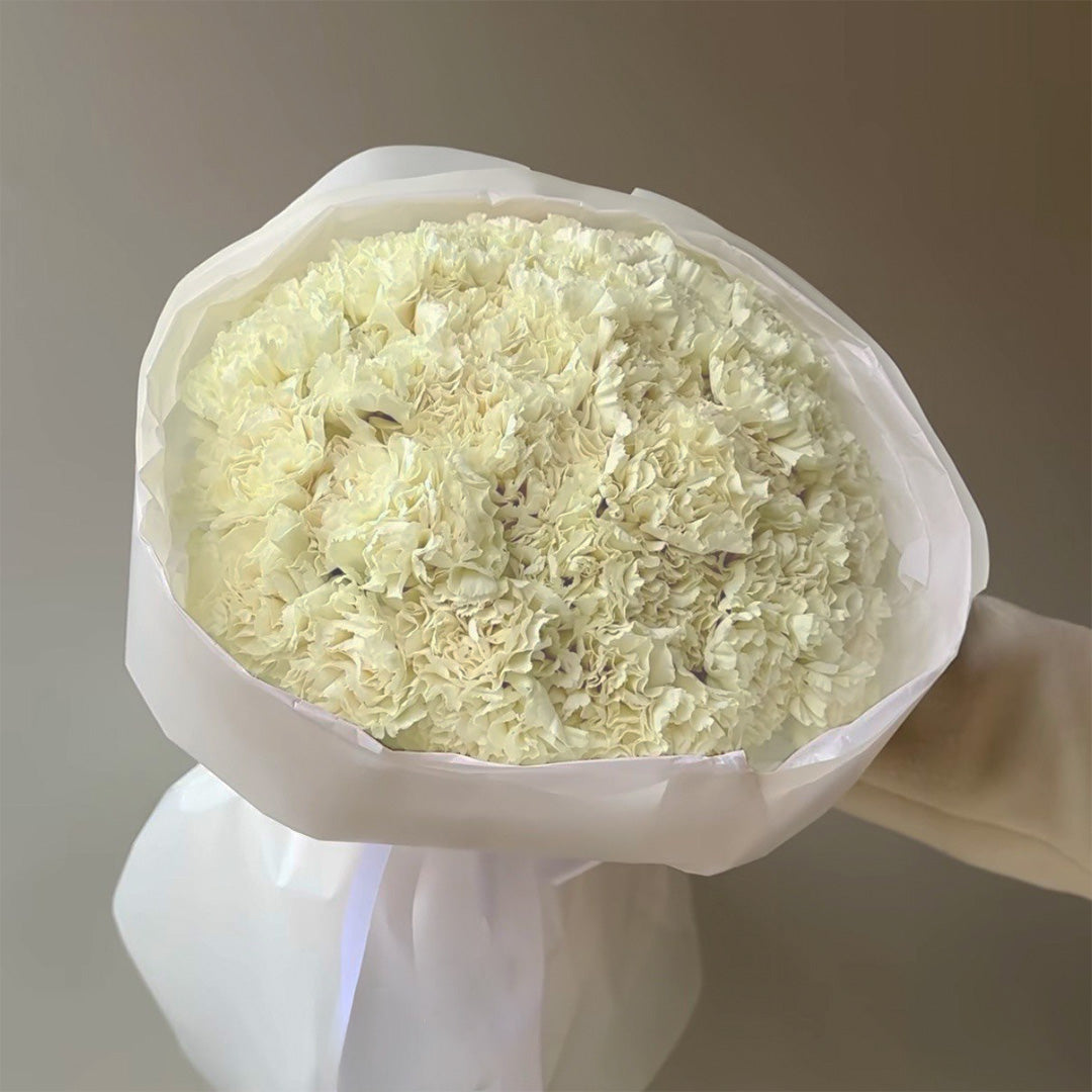 A bouquet of fresh white carnations arranged in a soft, rounded shape, wrapped in a delicate white paper. The flowers are tightly packed, displaying their lush texture and creating a full, elegant presentation.