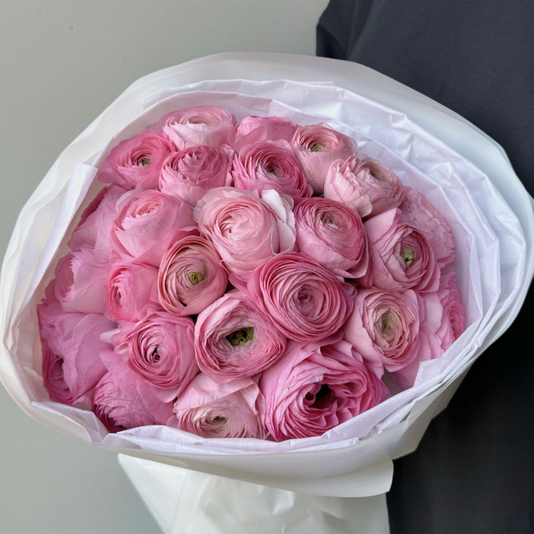 A beautiful bouquet of pink ranunculus flowers, carefully arranged with soft pink and white blooms. The flowers are tightly packed together in a neat, round shape, wrapped elegantly in white paper with a clean and classic appearance.