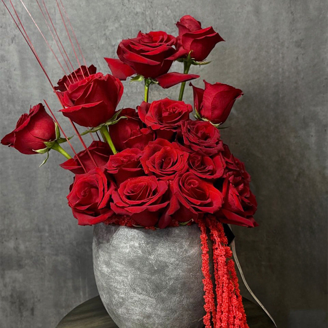 Vibrant red rose arrangement in a modern grey vase, complemented by cascading red tassels and delicate floral accents. A striking and romantic centerpiece, perfect for anniversaries, Valentine’s Day, or any special celebration.