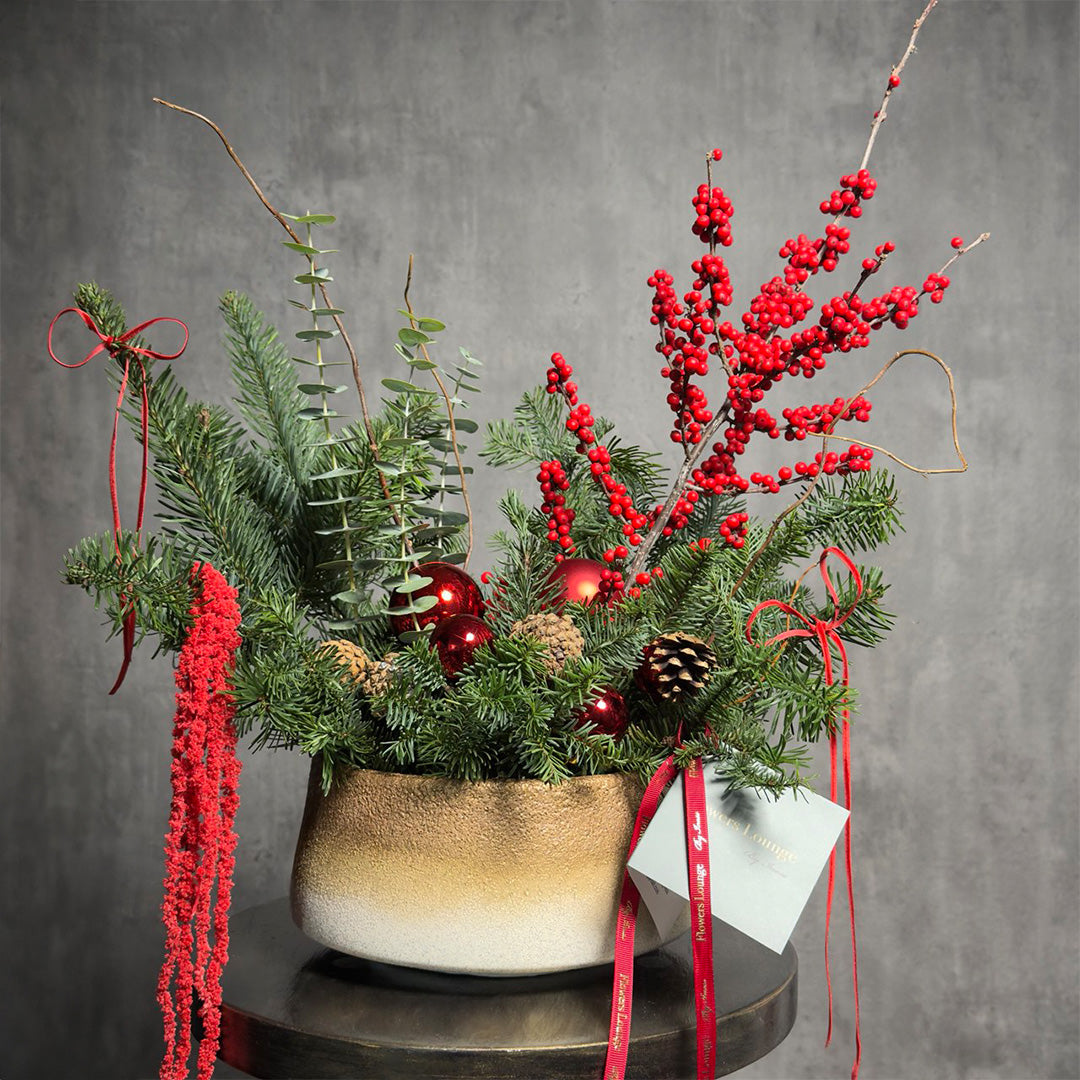 Holiday floral arrangement with red berries, pine, ornaments, and eucalyptus in a gold ceramic vase – Crimson Noel by Flowers Lounge.
