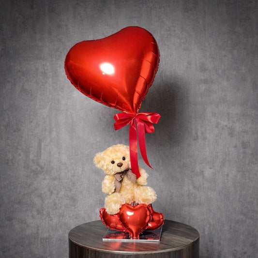 Teddy bear holding a red heart balloon with red ribbon, displayed on a clear base.