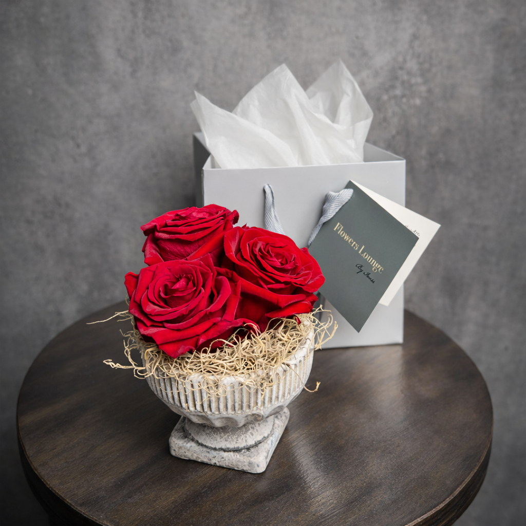 Three preserved red roses arranged in a decorative stone vase, with branded gift bag from Flowers Lounge by Iness.