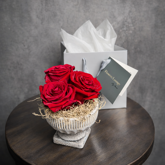 Three preserved red roses arranged in a decorative stone vase, with branded gift bag from Flowers Lounge by Iness.