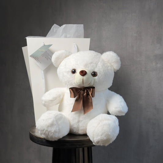 Plush white teddy bear with a satin brown bow, seated in front of a gift bag on a dark wood table – White Cuddle Gift