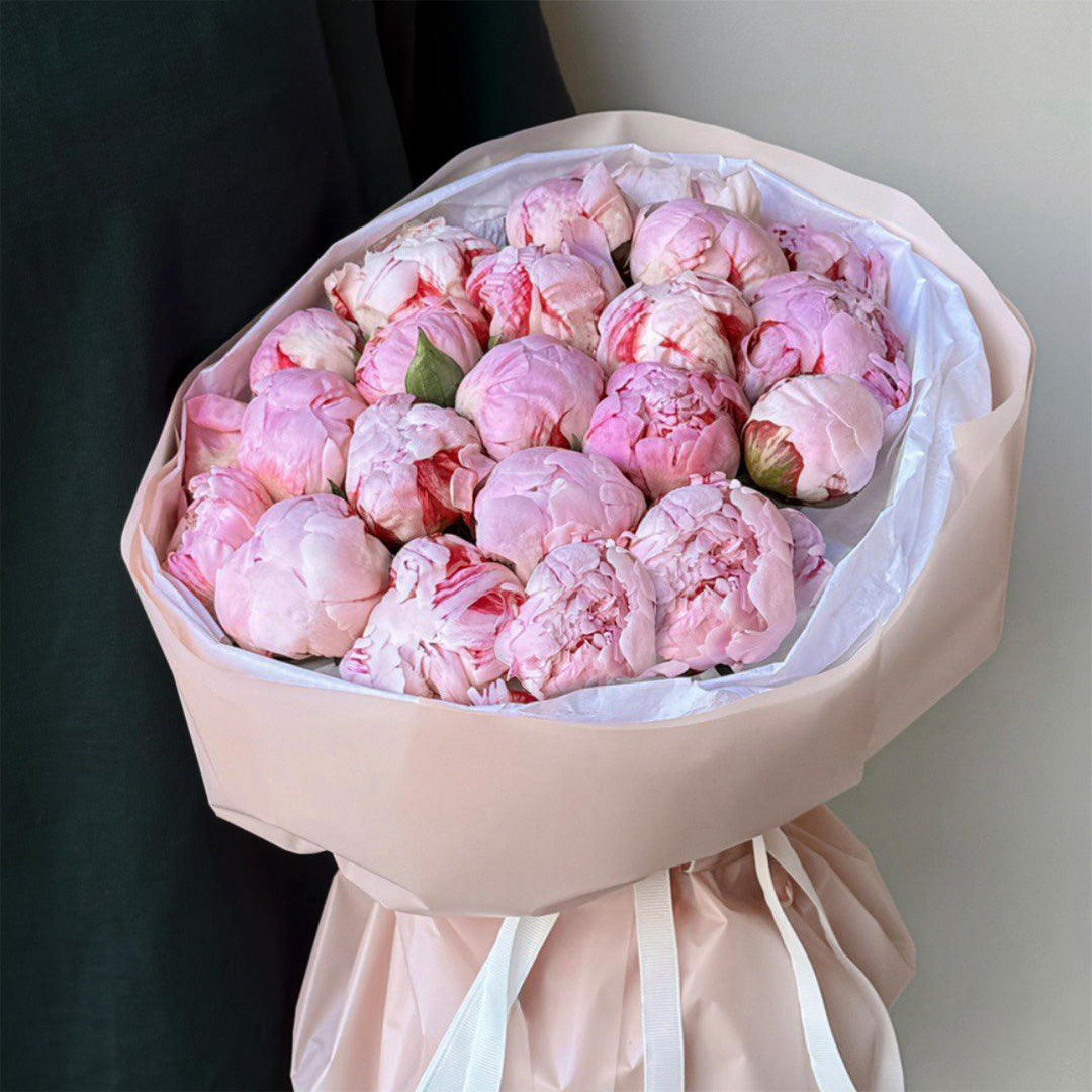 A stunning bouquet of soft pink peonies, arranged elegantly in a pale blush-colored wrapping. The flowers are freshly bloomed, with their petals in various shades of pink, creating a romantic and delicate feel. The bouquet is tied with a white ribbon, enhancing its charm.