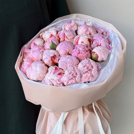 A stunning bouquet of soft pink peonies, arranged elegantly in a pale blush-colored wrapping. The flowers are freshly bloomed, with their petals in various shades of pink, creating a romantic and delicate feel. The bouquet is tied with a white ribbon, enhancing its charm.