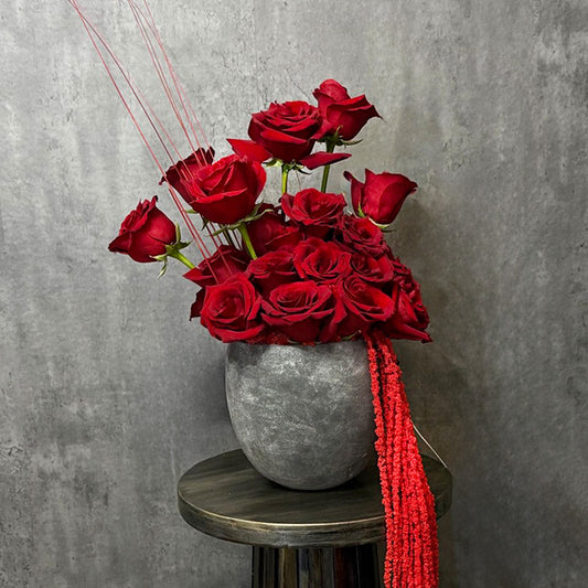 Luxurious bouquet of deep red roses in a modern grey vase, accented with delicate red strands and textured ribbons. A sophisticated floral arrangement perfect for expressing love and admiration, ideal for anniversaries, Valentine’s Day, or special celebrations