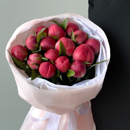 A bouquet of lush pink peonies tightly packed in a round shape, showcasing the elegant curvature of the buds that are just beginning to open. The peonies are complemented with dark green leaves, all wrapped in white paper with a delicate satin ribbon.
