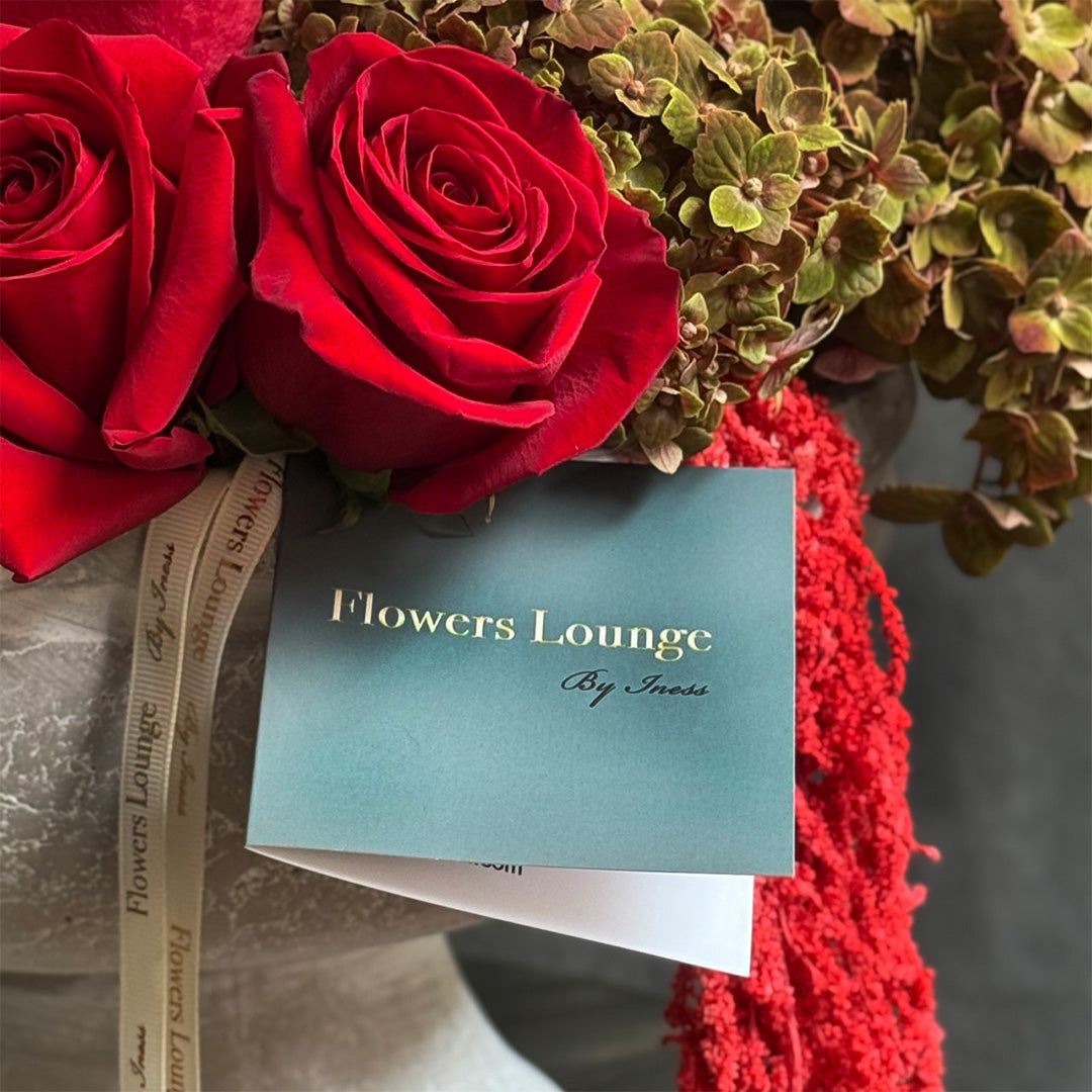 Close-up of two deep red roses with a luxurious ribbon featuring ‘Flowers Lounge by Iness,’ complemented by soft green foliage and a branded card. Perfect for romantic gestures, anniversaries, or special occasions, capturing elegance and timeless beauty.