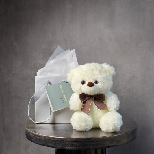 Cream-colored plush teddy bear with a brown bow, seated in front of a white gift bag with tissue paper, on a dark round table against a soft gray background.
